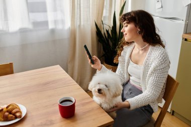 Genç bir kadın, kabarık tüylü bir köpek eşliğinde telefonuna bakarken sabah kahvesinin tadını çıkarıyor..