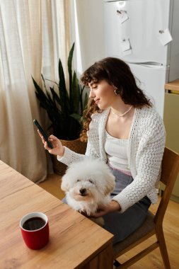 Evde dinlenen genç bir kadın kahvesini yudumluyor ve köpeğiyle birlikte akıllı telefonuyla etkileşime geçiyor..