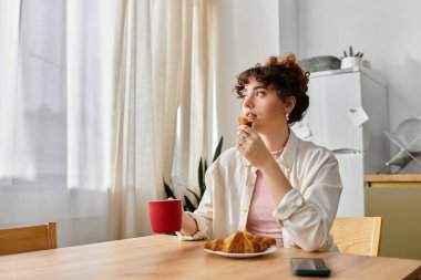 Genç bir kadın kahvesini yudumluyor ve bir kruvasan kemiriyor. Düşüncelere dalmış, güneşli bir masada..