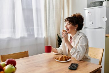 Genç bir kadın güneşli bir sabahta mutfak masasında kruvasanların ve kahvenin tadını çıkarırken gülümsüyor..