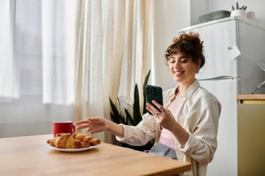 Genç bir kadın güneşli bir mutfak masasında oturuyor, pastalardan zevk alıyor ve telefonuyla meşgul oluyor..