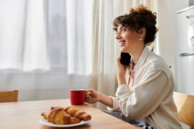 Genç bir kadın sabah güneşi altında telefonuyla sohbet ederken evinde çay yudumluyor..