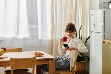 Genç bir kadın kahvesini yudumlarken gülümsüyor ve mutfak masasında telefonunu karıştırıyor..