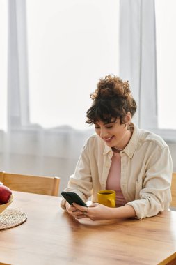 Neşeli bir kadın telefonuna gülümsüyor, evinde sıcak bir fincan kahve tutuyor..