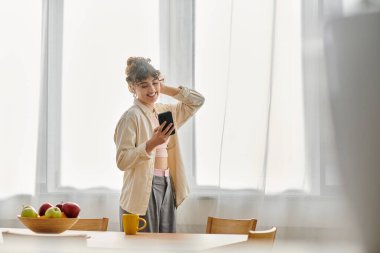 Neşeli genç bir kadın parlak bir mutfakta dururken akıllı telefonuna gülümsüyor..