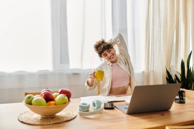 Relaxed young woman stretches while sipping tea at her cozy breakfast table in the morning sun clipart