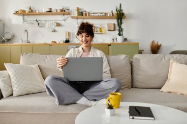 Genç bir kadın kanepede bacak bacak üstüne atarak oturuyor, keyifle şık bir odada laptopunu kullanıyor..