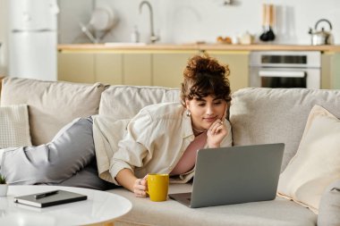 Genç bir kadın kanepede dizüstü bilgisayarıyla rahatlar ve sıcak bir içecek yudumlar..