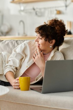 Genç bir kadın kanepede dinleniyor, laptopuyla meşgul ve sıcak bir içkinin tadını çıkarıyor..