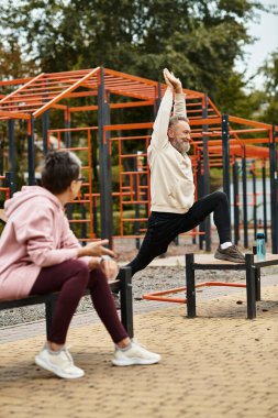 Bir çift açık hava spor salonunda eğleniyor, erkek esniyor ve kadın gülümsüyor..