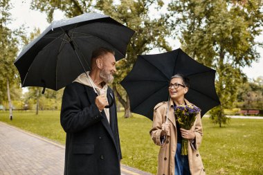 Sevgi dolu bir çift şemsiyelerle el ele yürür, canlı yeşil bir parkta gülümsemelerini paylaşırlar..