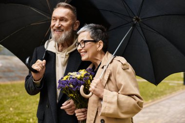 Sevgi dolu bir çift, yağmurlu bir günde şemsiye ve çiçek tutarak birlikte yürürken gülümser..