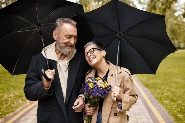 Bir çift, şemsiye ve çiçekleri tutarken gülücüklerini paylaşarak parkta keyifli bir yürüyüş yapıyor..