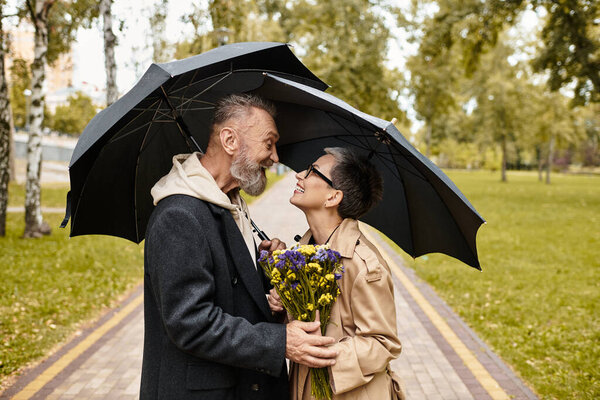 A couple shares joyful smiles and a bouquet, standing close under an umbrella.