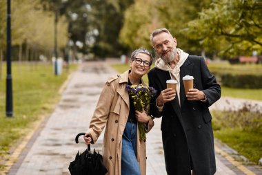 Gülümseyen bir çift, elinde kahve fincanları ve çiçeklerle parkta neşeli bir anı paylaşır..
