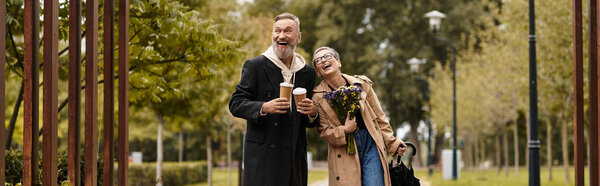 A joyful couple enjoys warm beverages while strolling through a peaceful park filled with greenery.