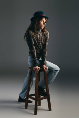 A handsome man with long hair models unique attire while sitting on a wooden stool in soft lighting. clipart