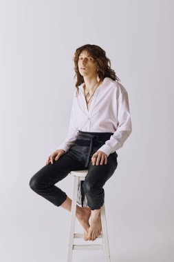 Uzun saçlı yakışıklı genç bir adam taburede çıplak ayakla oturur, rahat bir zarafet yayar..