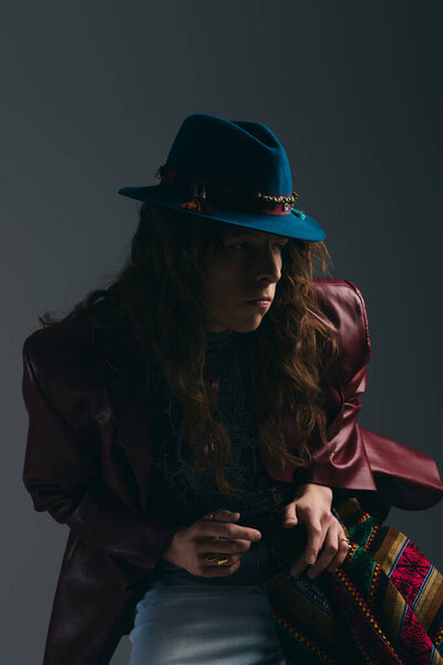 Модный молодой человек с длинными волосами и красочной шляпой творчески позирует в современной студии.