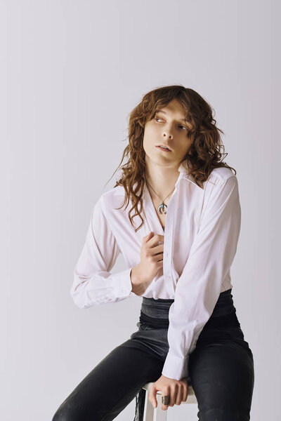 A handsome young man exudes charm while seated, dressed in a sleek button up and fitted slacks.