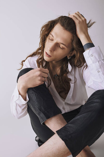 Красивый молодой человек с длинными волосами сидит небрежно, создавая расслабленную, но стильную атмосферу.