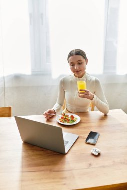Genç bir kadın evde dizüstü bilgisayarıyla uğraşırken canlı bir salata kahvaltısının tadını çıkarıyor..