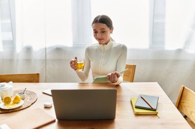 Genç bir kadın elinde dizüstü bilgisayarla mutfağında oturmuş, çayını yudumluyor ve işi için notları gözden geçiriyor..
