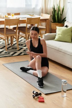 Siyah spor kıyafetli fit bir kadın şık bir dairede egzersiz yaparken telefonunu kontrol ediyor..