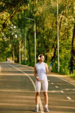 Genç bir atletik kadın su şişesiyle gülüyor, yemyeşil bir parkta mola veriyor..
