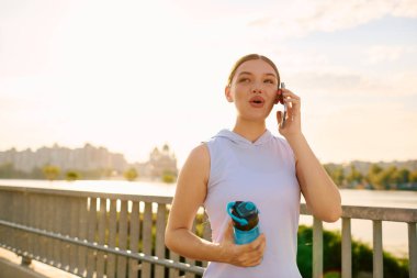 A young beautiful athlete engages in a cheerful call while jogging alongside a river at dusk. clipart