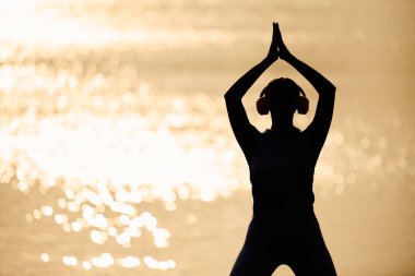 Genç kadın gün batımında su kenarında yoga yapıyor, huzur ve odaklanma yayıyor..