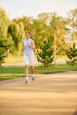 Parkta güneşli bir günün sıcaklığının tadını çıkararak, neşeli bir şekilde yolda koşan sportif bir kadın..