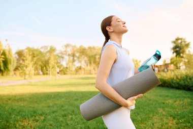 Güzel bir atletik kadın, canlı bir parkta gülümserken yoga minderi ve su şişesi taşıyor..