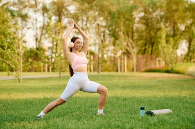 Genç, atletik bir kadın canlı bir parkta yumuşak çimenler üzerinde yoga pratiği yapıyor..