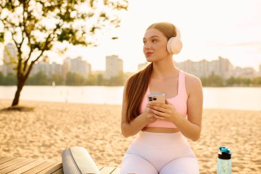 Spor kıyafetleri içinde genç bir kadın sahilde dinleniyor, suyu yudumluyor ve müzik dinliyor..