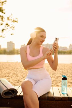 Sportif bir kadın göl kenarındaki bankta dinleniyor, güneş batarken telefonunun keyfini çıkarıyor..