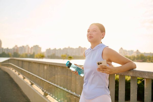 Beautiful young woman pauses to enjoy a drink on a bridge overlooking the city at dusk.