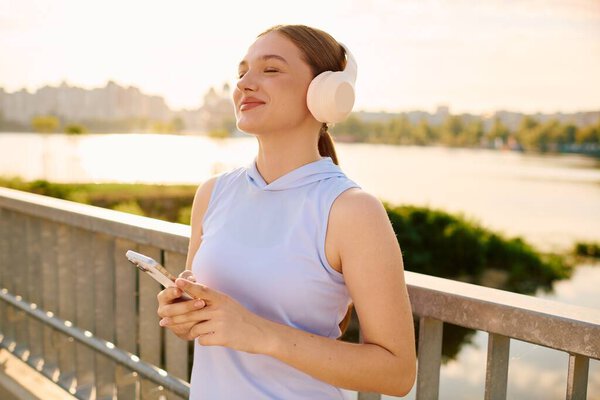 A young woman with headphones relaxes on a bridge, enjoying the sunset by the water.