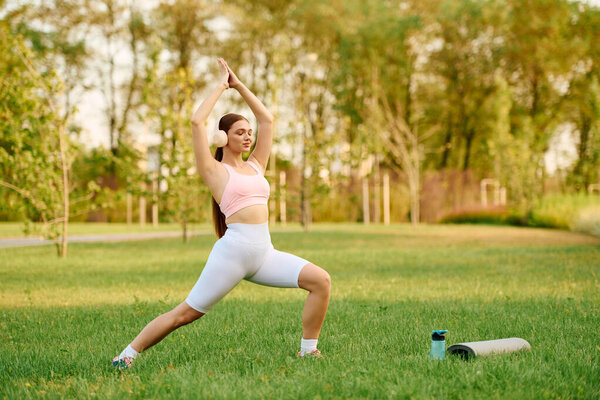 A young, athletic woman is gracefully executing a yoga practice on soft grass in a vibrant park.