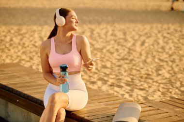 Genç bir kadın kumsalda gülümsüyor, telefonuna bakarken kulaklıkla rahatlıyor..