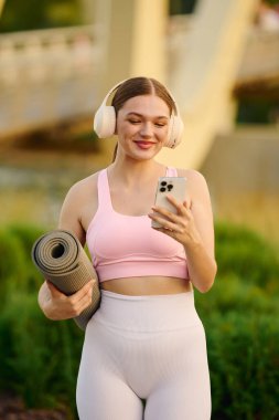Formda bir kadın, elinde yoga minderiyle parlak bir parkta duruyor ve telefonunu kontrol ediyor..