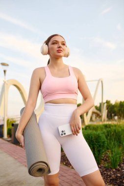 Dışarıda yoga minderi ve kulaklıklarıyla antrenman için hazır bekleyen genç bir kadın..