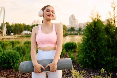 Aktif kadın, güneş batarken yoga minderi ve kulaklıklarla sakin bir parkta dinleniyor..
