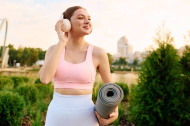 Spor kıyafetli neşeli genç kadın şehir parkında yemyeşil bir yoga için hazırlanıyor..