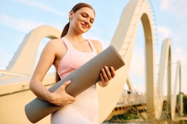 Enerjik genç bir kadın, modern köprü zeminli sakin bir açık havada yoga minderi tutuyor..