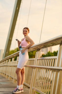 Atletik giyinen genç bir kadın, güneş batarken bir köprüde durup gülümsüyor ve telefonunu tutuyor..