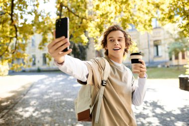 Sırt çantalı genç adam elinde kahve fincanıyla selfie çekerken geniş bir gülümseme sergiliyor..