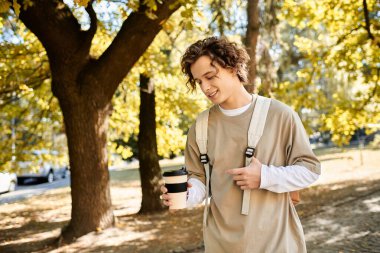 Kıvırcık saçlı yakışıklı bir öğrenci, parlak sonbahar yaprakları ve güneş ışığıyla çevrili kahvesini yudumluyor..