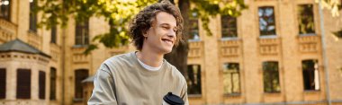 Young man smiles while sipping coffee, surrounded by autumn leaves in an elegant courtyard. clipart