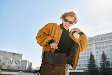 Young man with curly hair in a stylish jacket gets coffee and a notebook from his bag in the sun. clipart